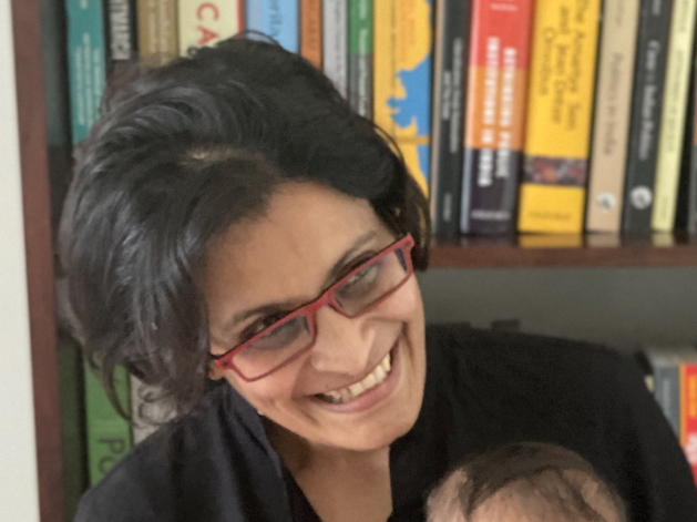 This is an image of Dr. Sai Balakrishnan holding her daughter. She is sitting in front of a bookcase, while smiling.
